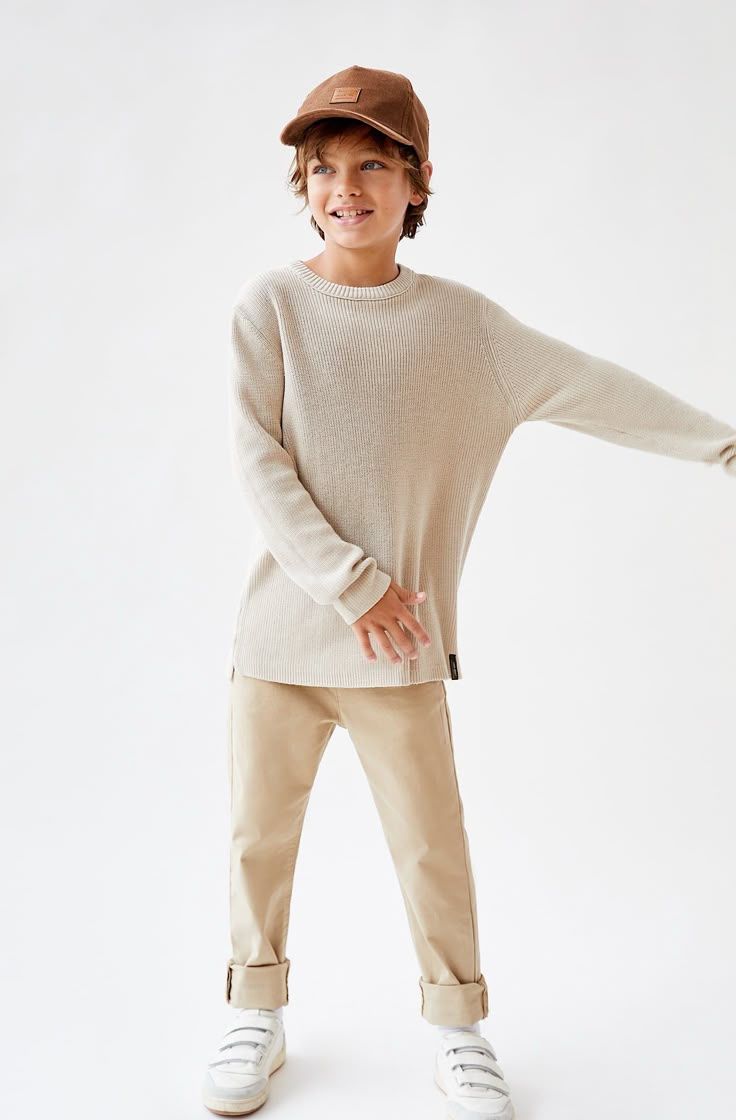 Boy wearing a beige knit sweater, tan chinos, white sneakers, and a brown cap in a casual pose.