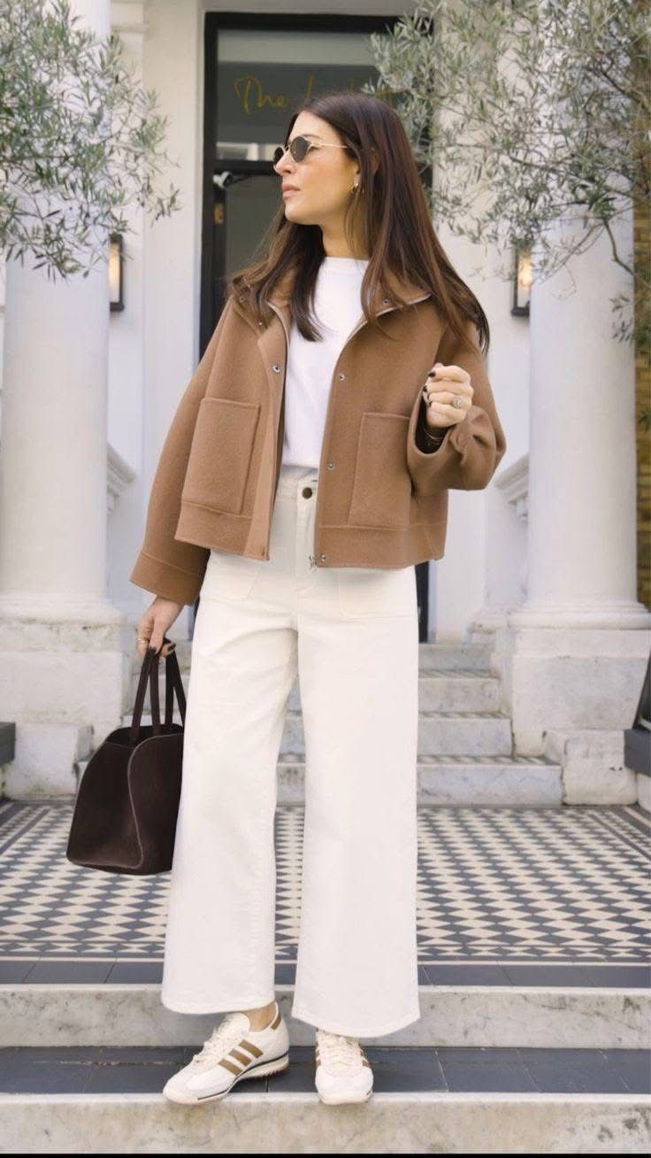Woman wearing a camel wool jacket, white top, cream wide-leg pants, and gold sneakers holding a dark bag.
