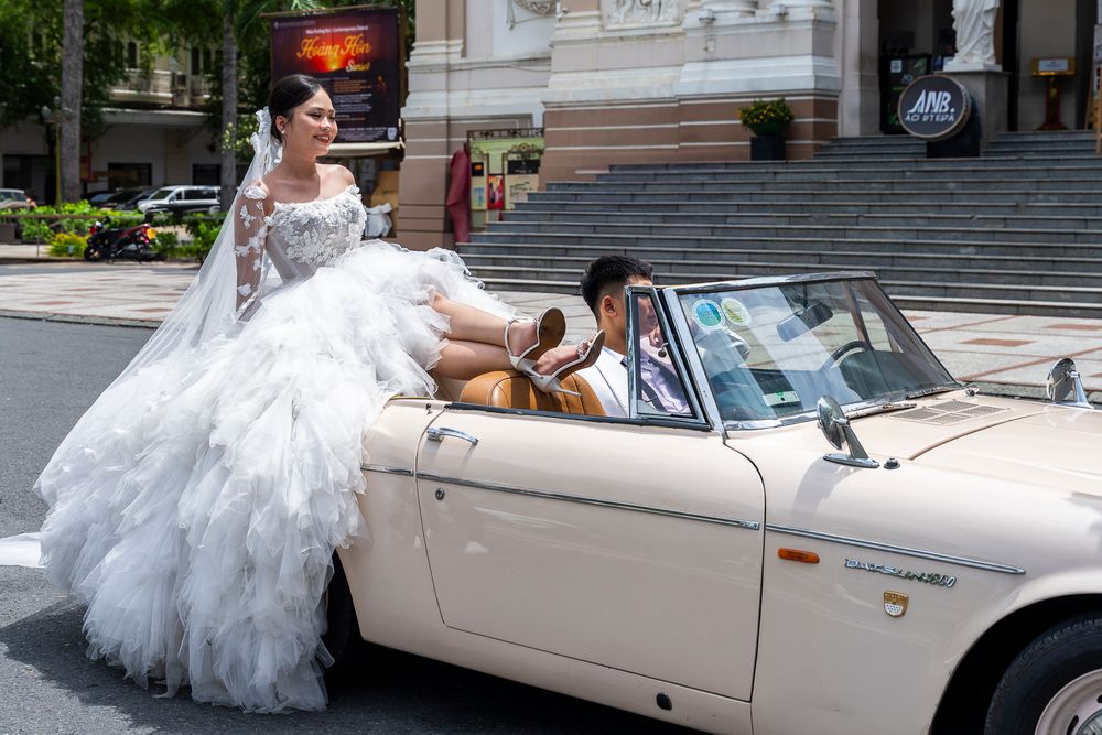 Bride in elegant white ruffled gown poses beside a classic white convertible on a city street.
