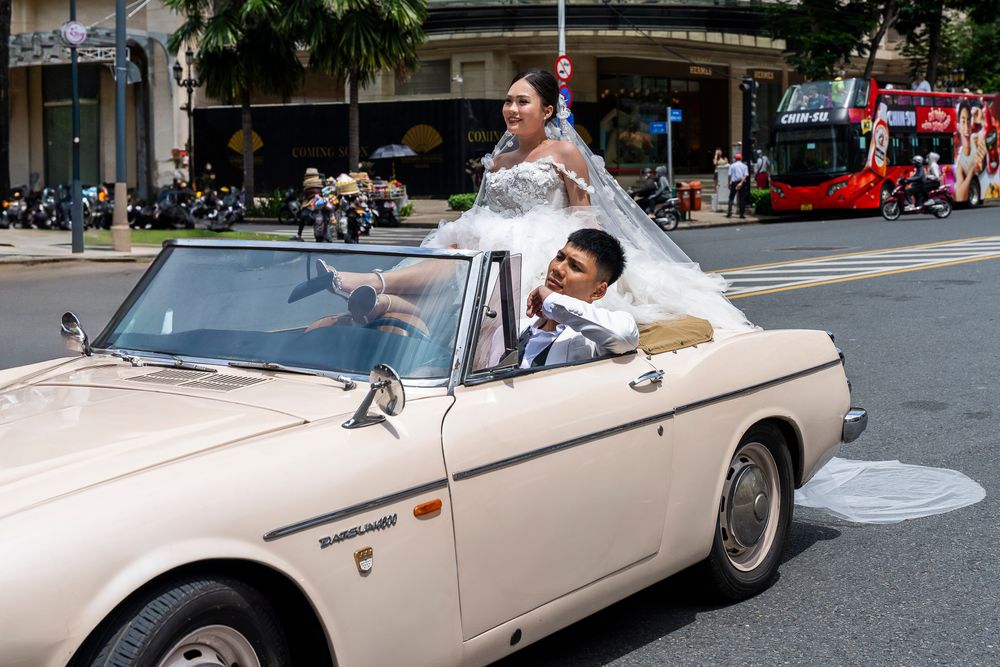 Couple in wedding attire rides in vintage cream convertible car through city street.