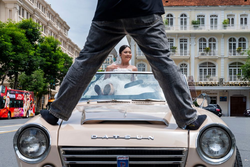 Photographer stands on hood of vintage convertible car capturing bride inside during wedding shoot.