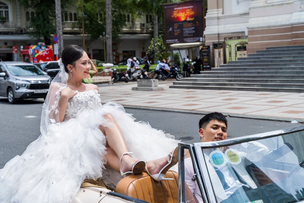 Bride in white gown sits on convertible classic car with groom during outdoor wedding photo shoot.