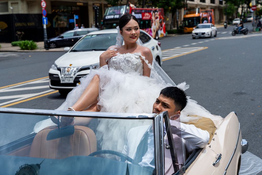 Bride in strapless white gown and groom in white suit pose in open-top classic car on city street.