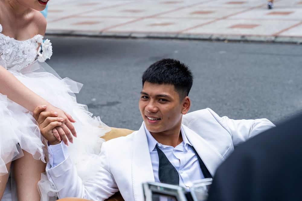 Smiling groom in white suit holds bride's hand during outdoor wedding photo session.