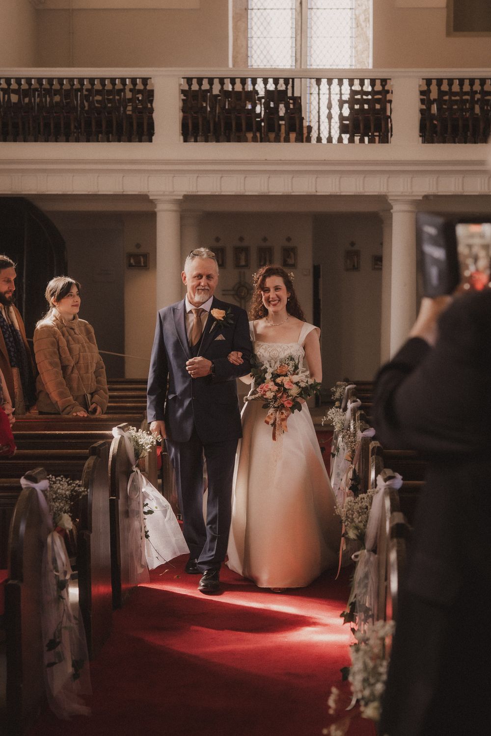 Bride walks down red carpet aisle with her father in a historic church, holding a floral bouquet on her wedding day.