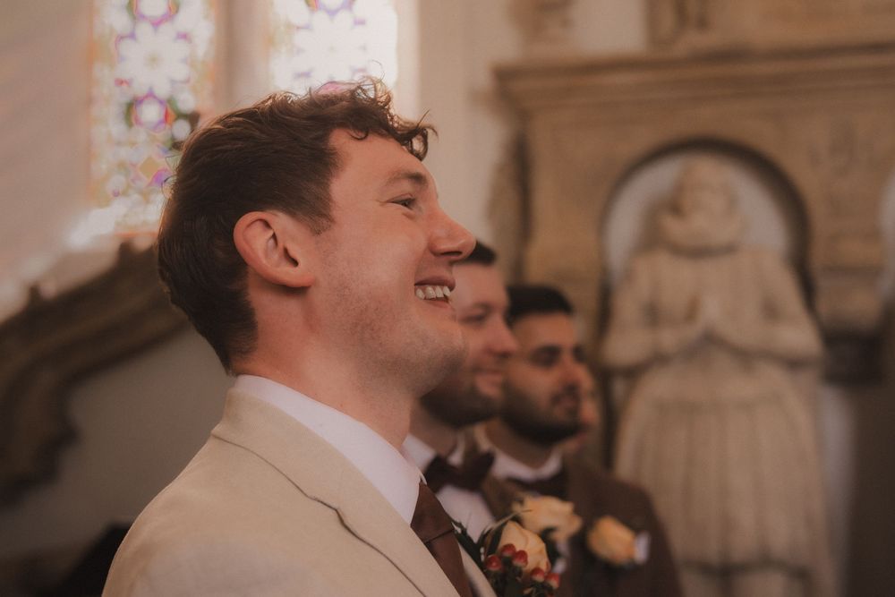 Groom smiling joyfully at the altar in a beige suit with floral boutonniere inside a historic church.