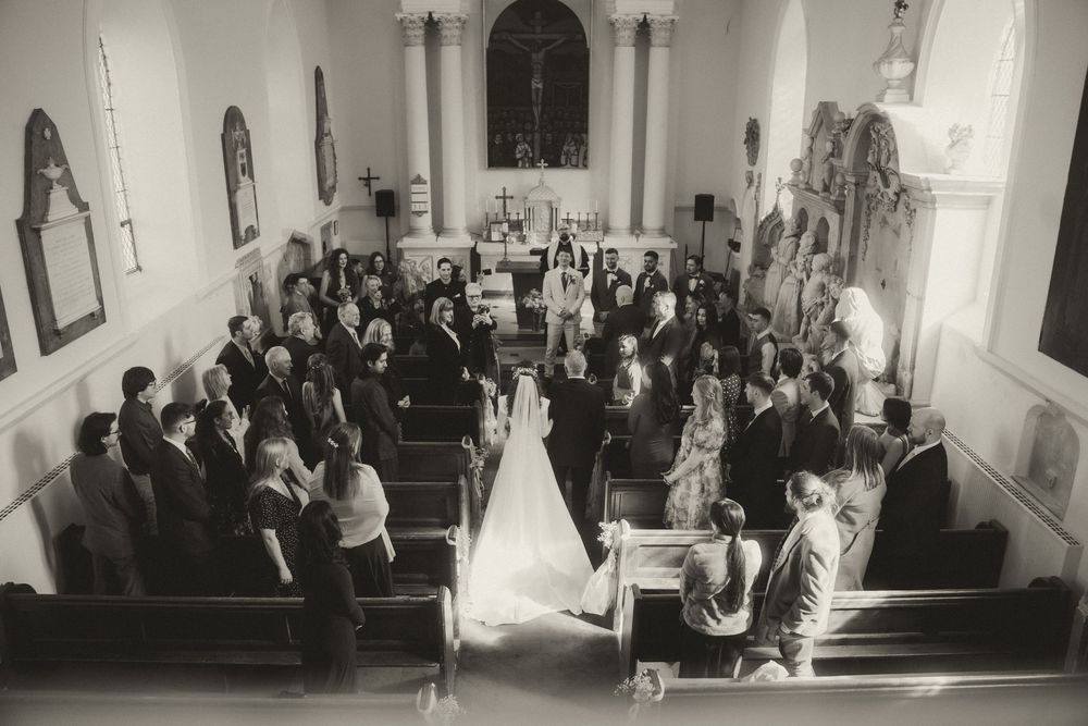 Black and white aerial view of a wedding ceremony inside a small church with guests seated in pews.