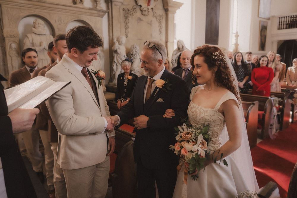 Bride and groom exchange vows at elegant church wedding ceremony, with father of bride and guests in background.
