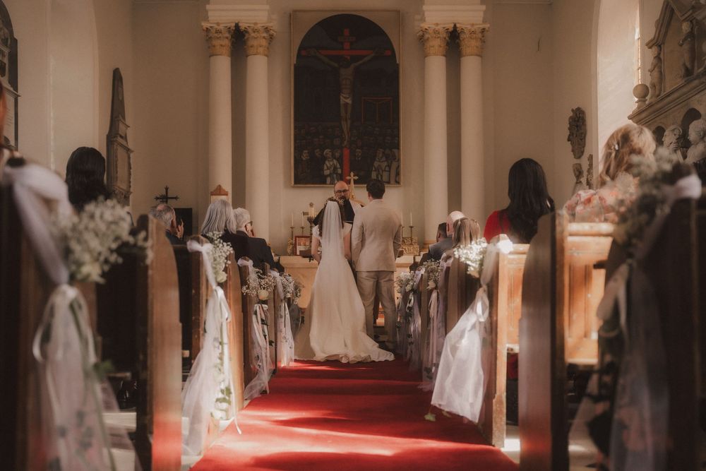A bride and groom stand at the altar of an elegant church with red carpet aisle and white floral pew decorations.
