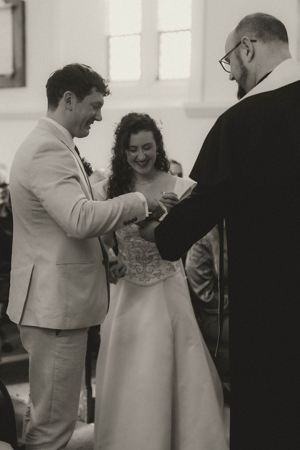 Black and white photo of a smiling bride and groom exchanging rings at the altar during a church wedding ceremony.