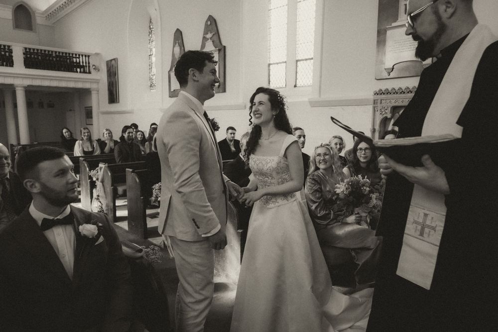 Black and white wedding ceremony photo of bride and groom exchanging vows before officiant in a church.