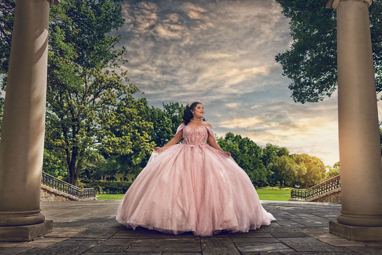 A girl in a shimmering blush pink Quinceañera gown and tiara poses between stone pillars in an outdoor park at sunset.