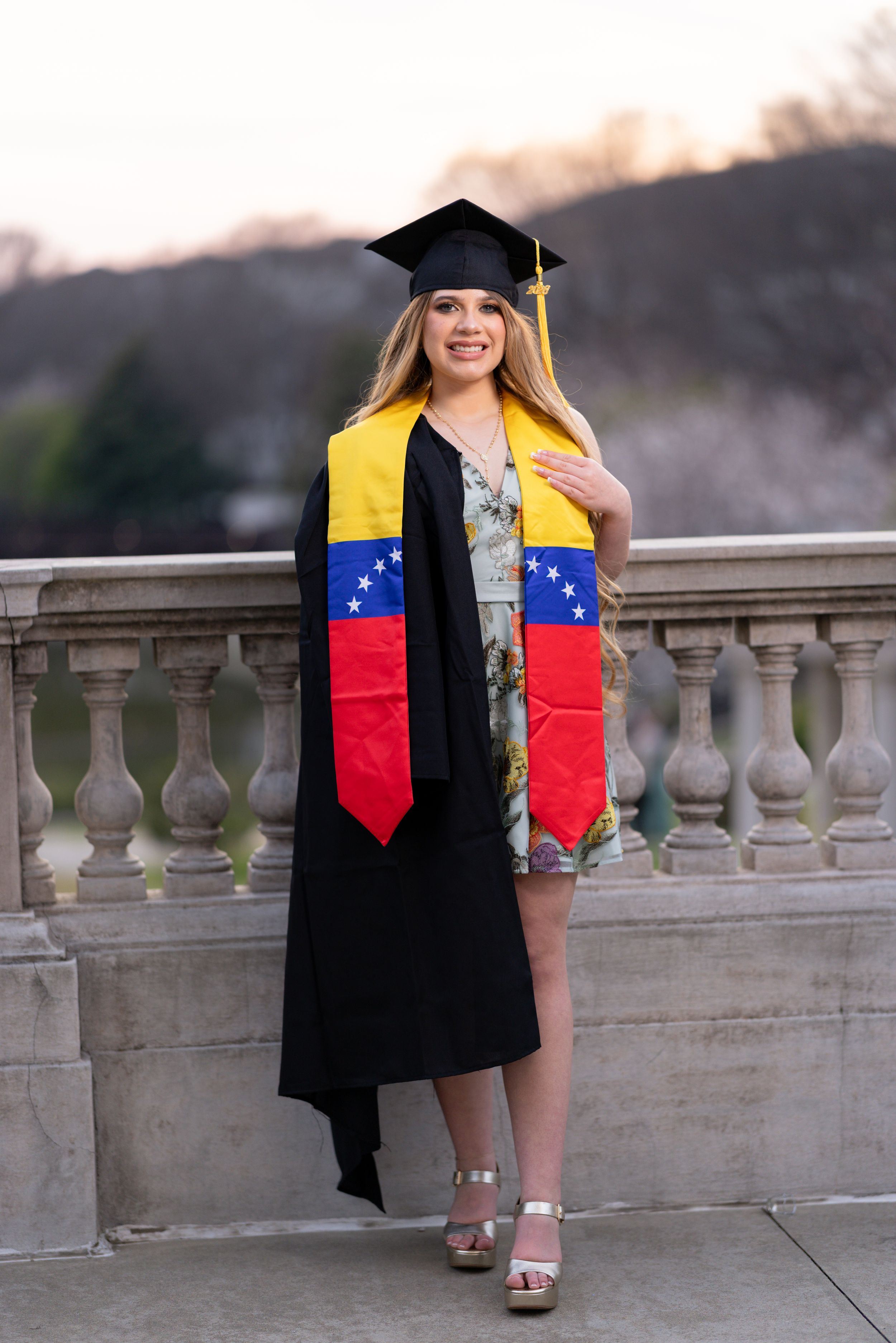 Smiling female graduate in black cap and gown wearing Venezuelan flag sash, posing outdoors near stone balustrade.