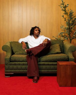 A young man with dreadlocks sits relaxed on a green sofa, wearing white shirt and burgundy pants on red carpet.
