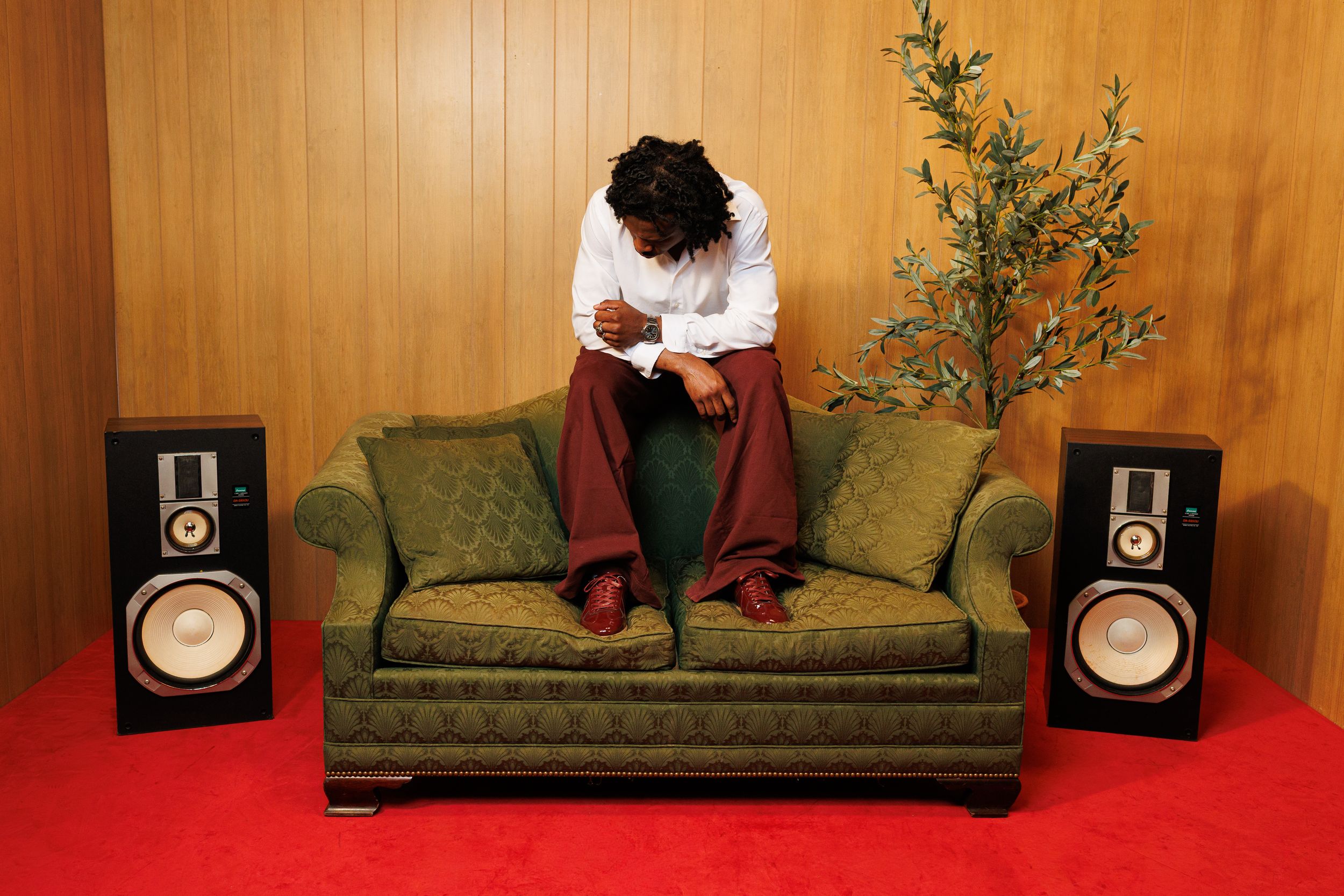 Man sitting on green couch between two speakers with a plant nearby on red carpet floor.