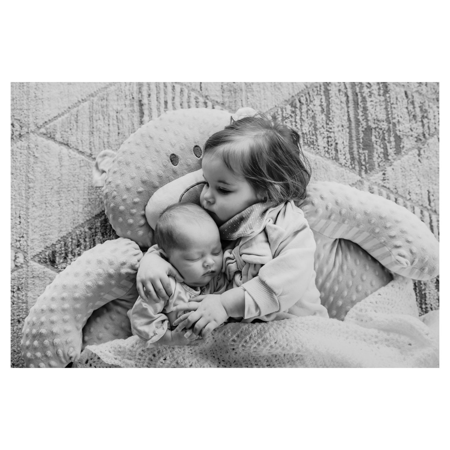 A black and white photo showing tender siblings embracing while sitting on a plush surface together.