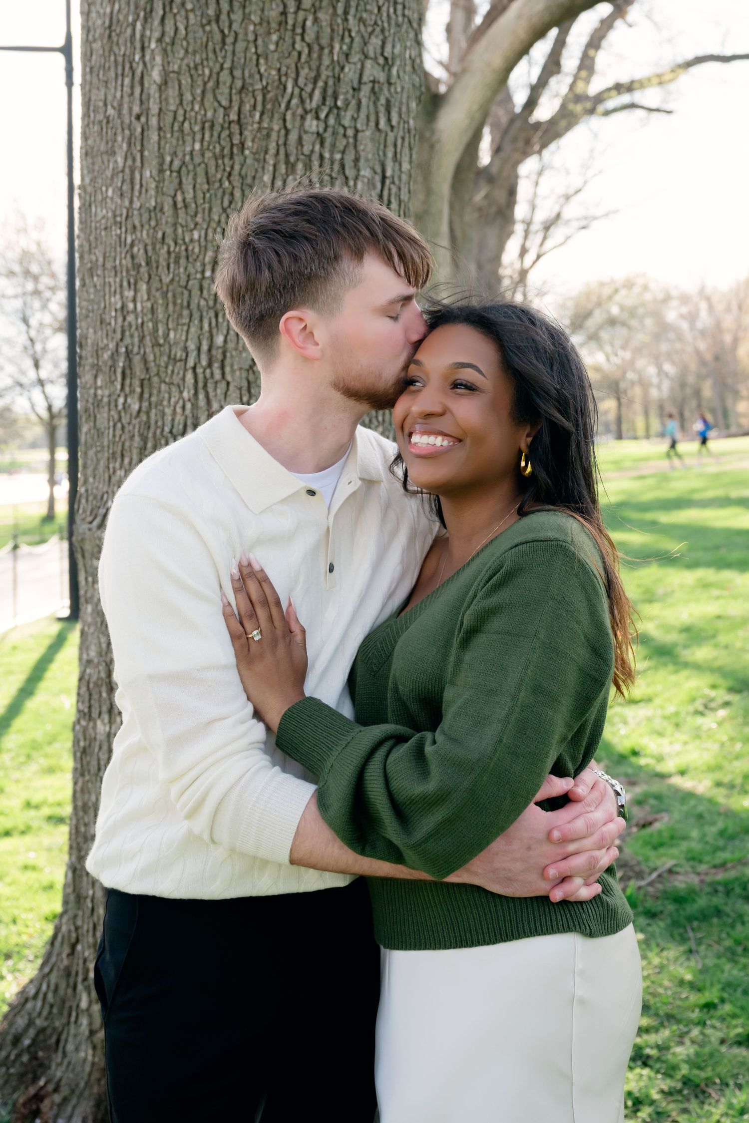 A joyful interracial couple embraces near a tree in a park, the man kissing the woman's cheek as she smiles.