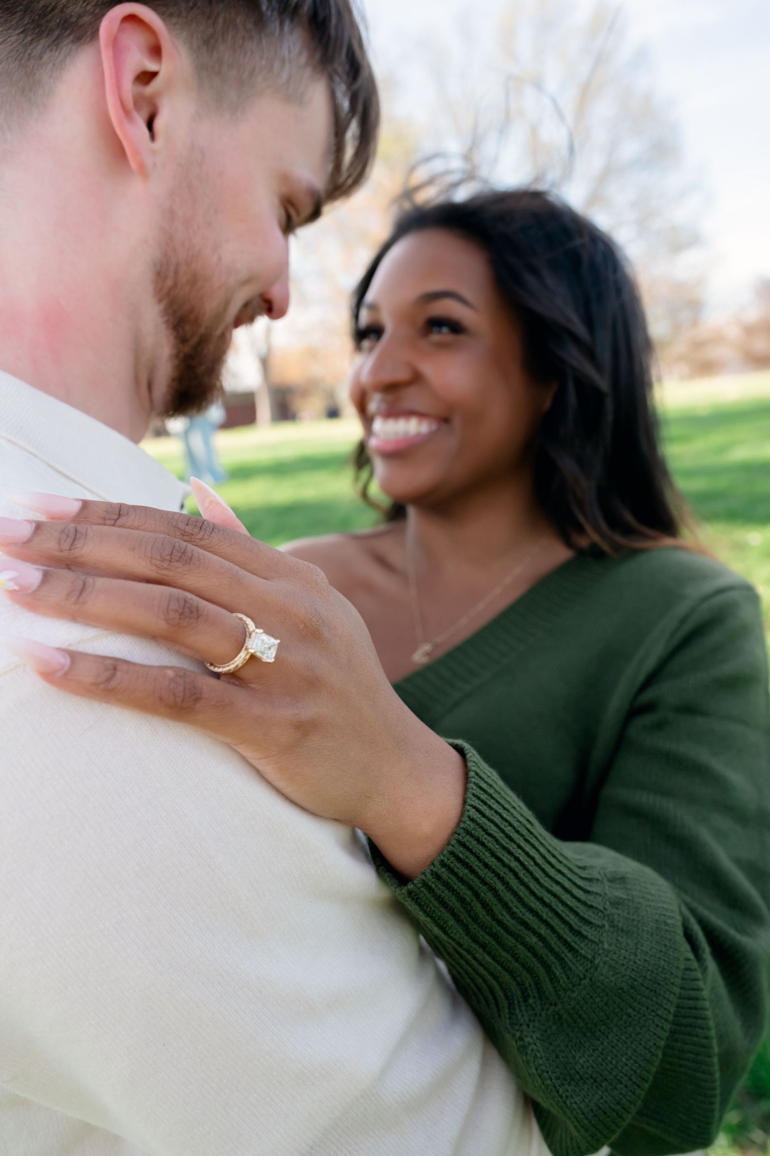 A smiling Black woman in a green sweater shows off her engagement ring while embracing her partner outdoors in a park.