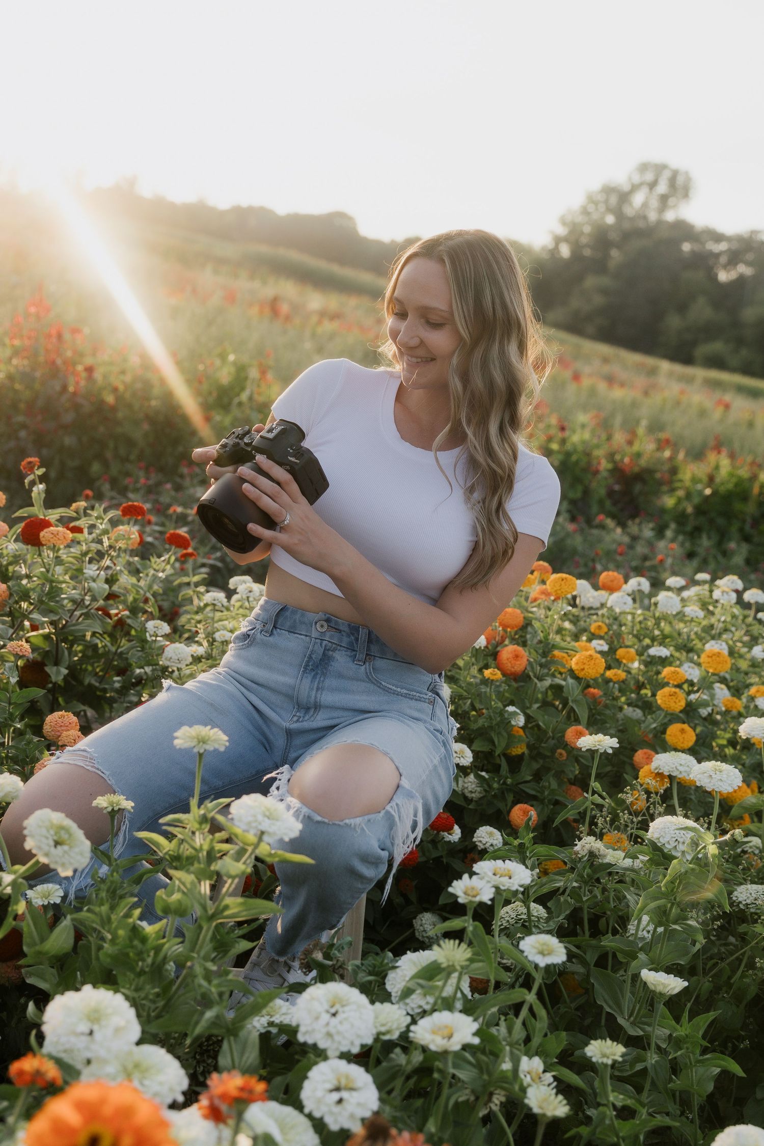 Blonde female photographer smiling while reviewing photos on camera in colorful flower field at golden hour.
