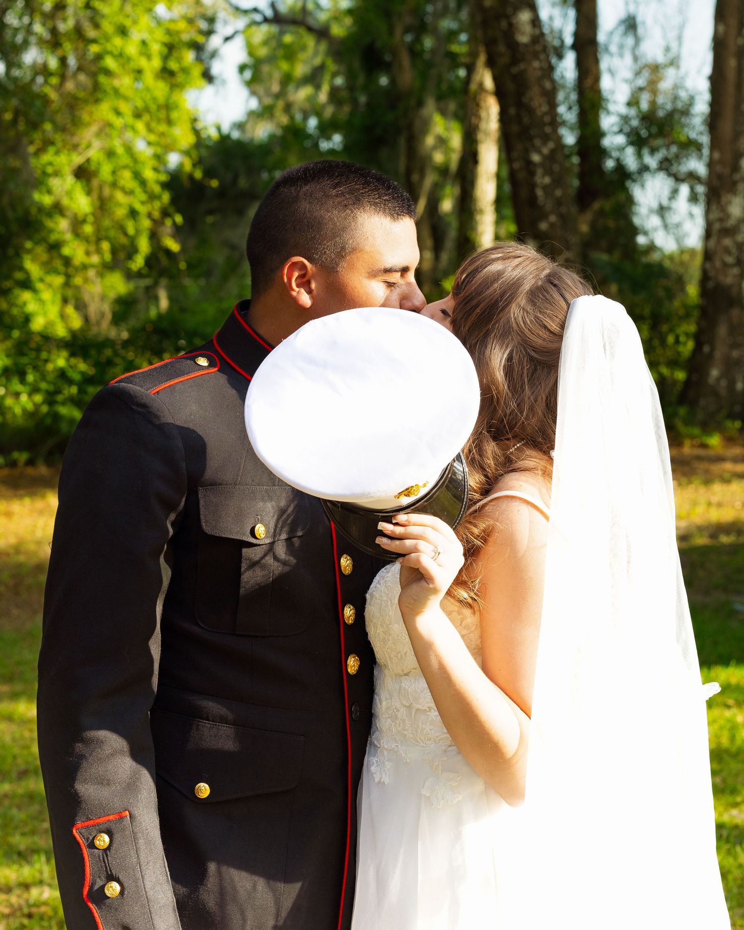 Marine in dress uniform stands behind bride on tree swing in sunlit outdoor wedding photo.