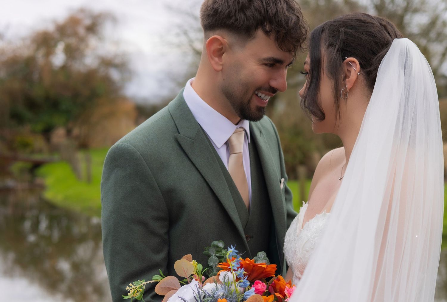 Bride and groom share an intimate moment outdoors, she holds a vibrant bouquet, he wears a sage green suit.