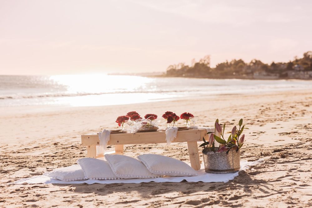 Romantic beach setup with red flowers on a wooden table, white cushions on sand, at golden sunset by the ocean.