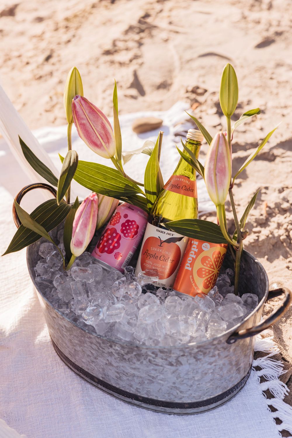 Metal bucket filled with ice, colorful beverage cans, a green bottle, and pink lily flowers on a sandy beach.