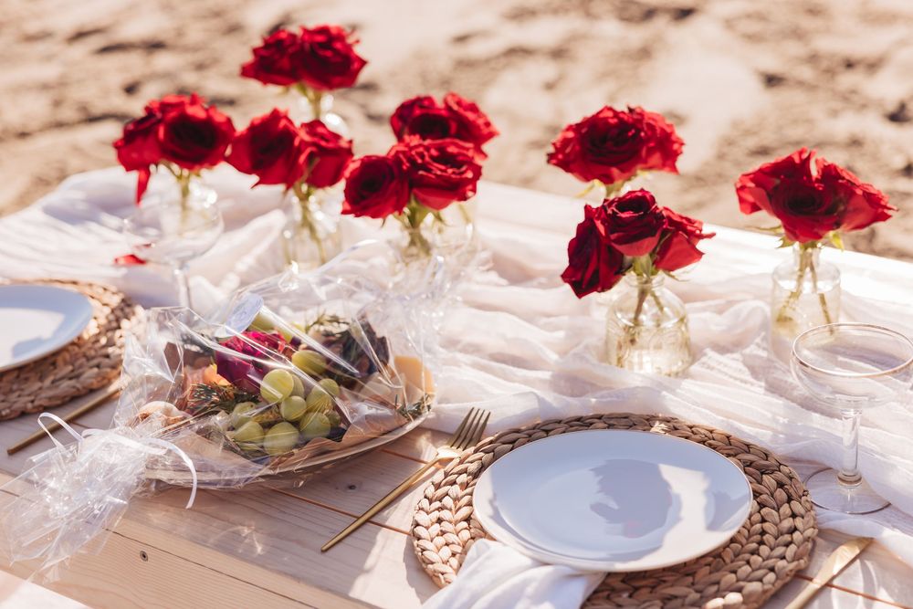 Romantic beach picnic setup with red roses in vases, white linen, wicker placemats, and elegant plates on sandy shore.