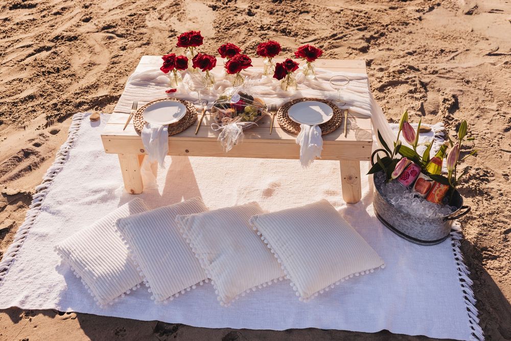 Romantic beach picnic setup with low wooden table, red roses, plates, and champagne bucket on white blanket with pillows.