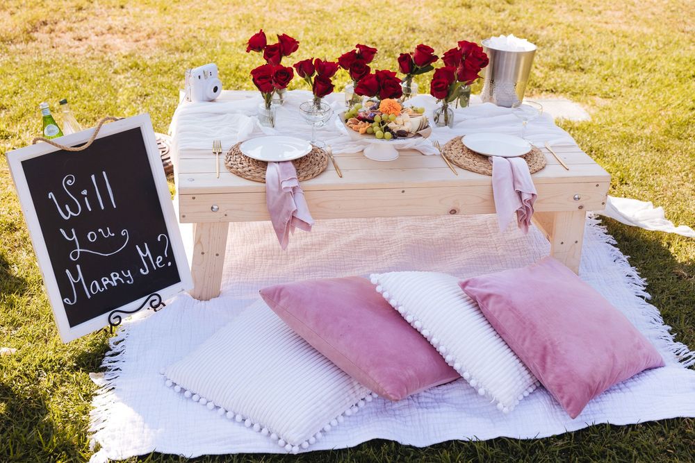 Romantic outdoor picnic setup with wooden table, red roses, pink pillows, and a Will You Marry Me chalkboard sign.