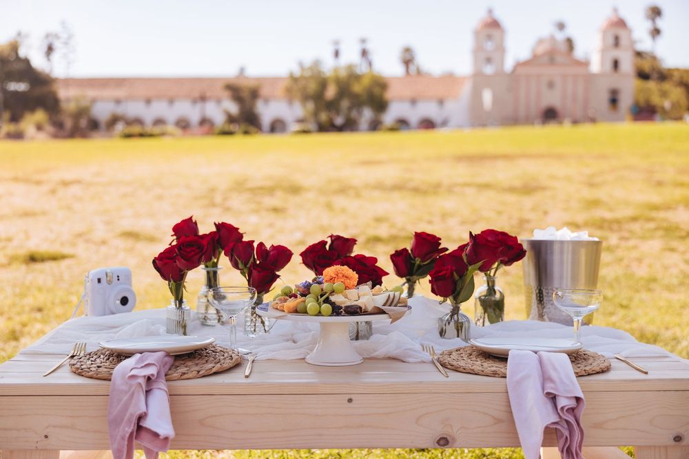 Outdoor picnic table with red roses, fruit, and place settings on grass near a white mission building.