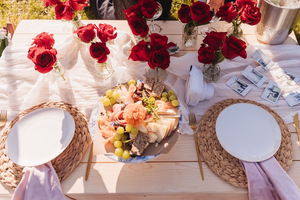 Elegant outdoor table setting with red roses, wicker placemats, white plates, and a charcuterie board with grapes and meats.