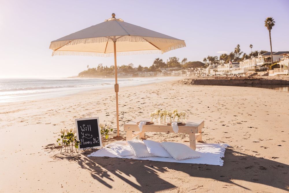 Romantic beach picnic setup with white umbrella, low table, and chalkboard sign on sandy California coastline.