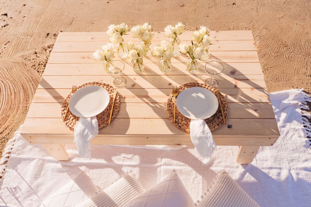 Romantic beach picnic setup with white plates, wicker chargers, white flowers, and a wooden table on sandy shore.