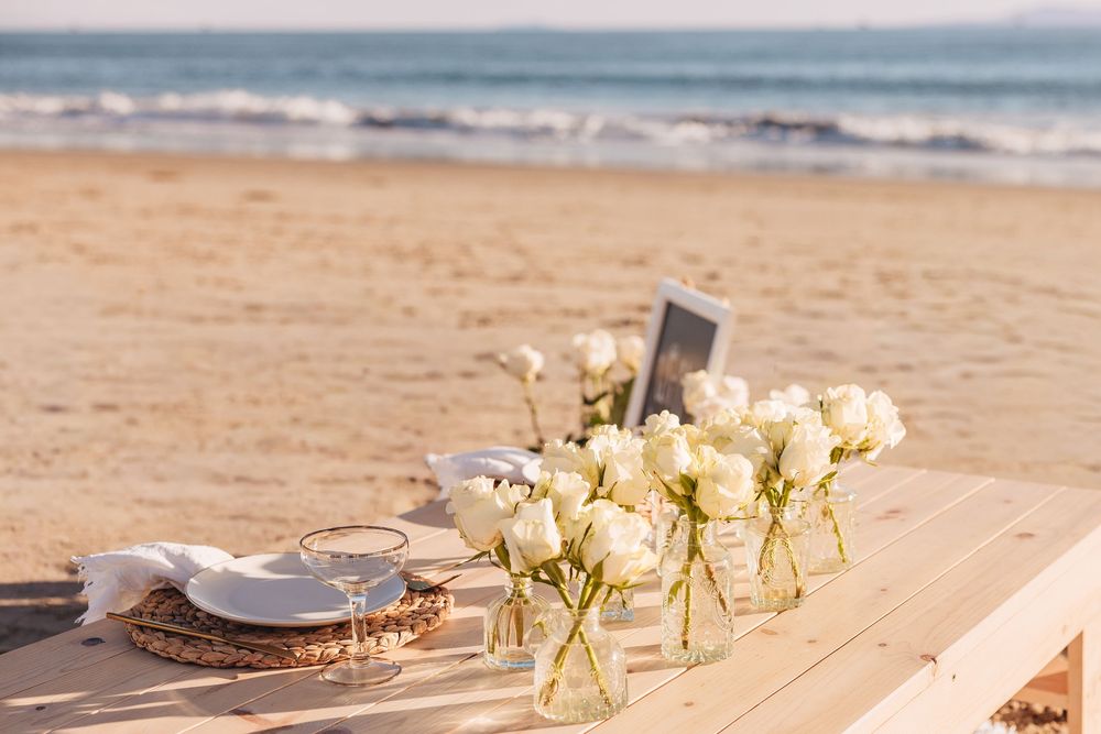 Romantic beach table setting with white roses in glass vases, wine glasses, and a chalkboard sign on sandy shore.