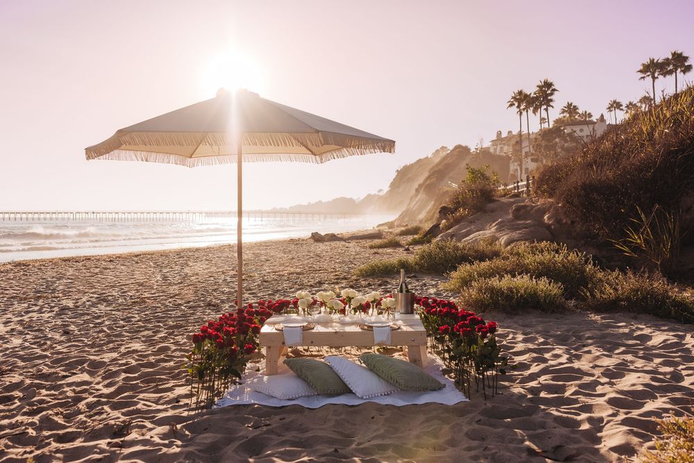 Romantic beach dinner setup with red roses, white table, and striped umbrella at golden sunset by the ocean.