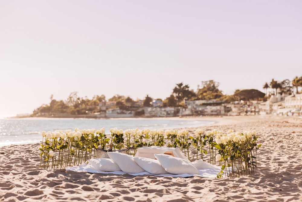 Beach wedding ceremony setup with white cushions and floral arrangements on sandy shore with ocean view.