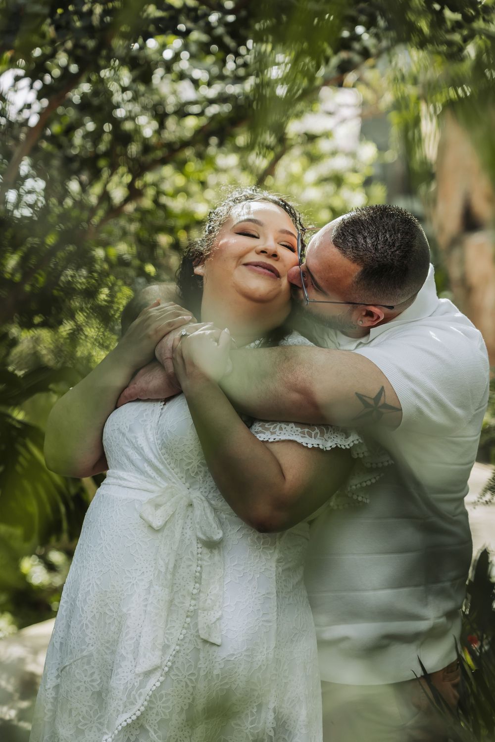 Groom kissing bride on cheek in lush garden setting, bride smiling joyfully in white lace dress.