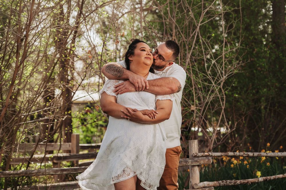 Couple embracing outdoors near wooden fence surrounded by lush green trees, woman in white dress with man in white shirt.