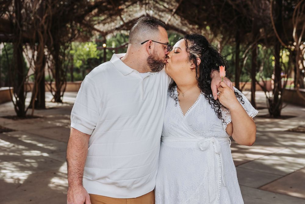 Couple sharing a kiss outdoors under a tree-covered pergola in white outfits.