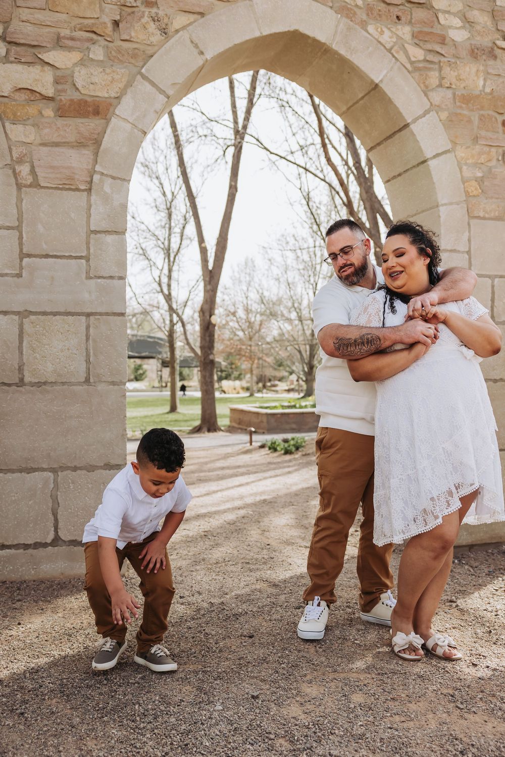 A family poses under a stone arch outdoors, with parents embracing and a young boy bending down on a gravel path.