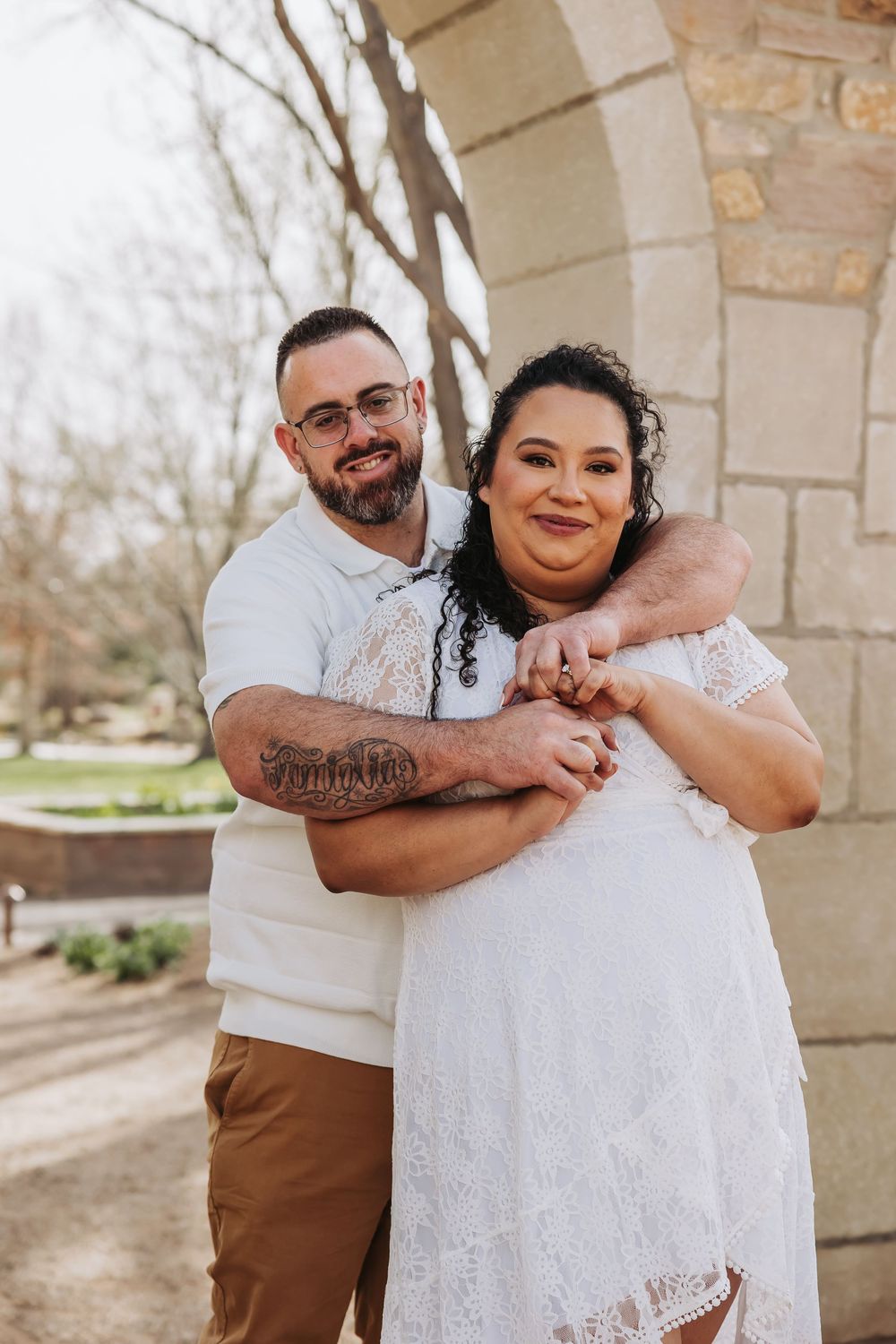 Couple embracing outdoors near stone arch, man with tattoos hugging woman in white lace dress, smiling warmly.