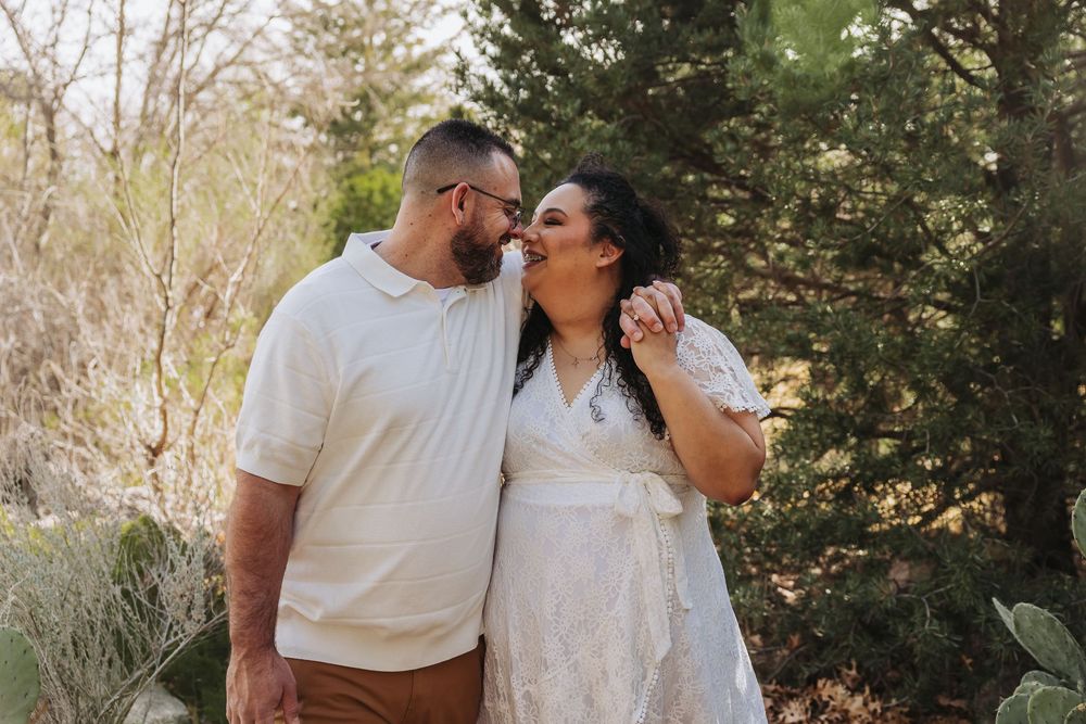Couple sharing a kiss outdoors surrounded by lush greenery, woman in white lace dress and man in cream polo shirt.