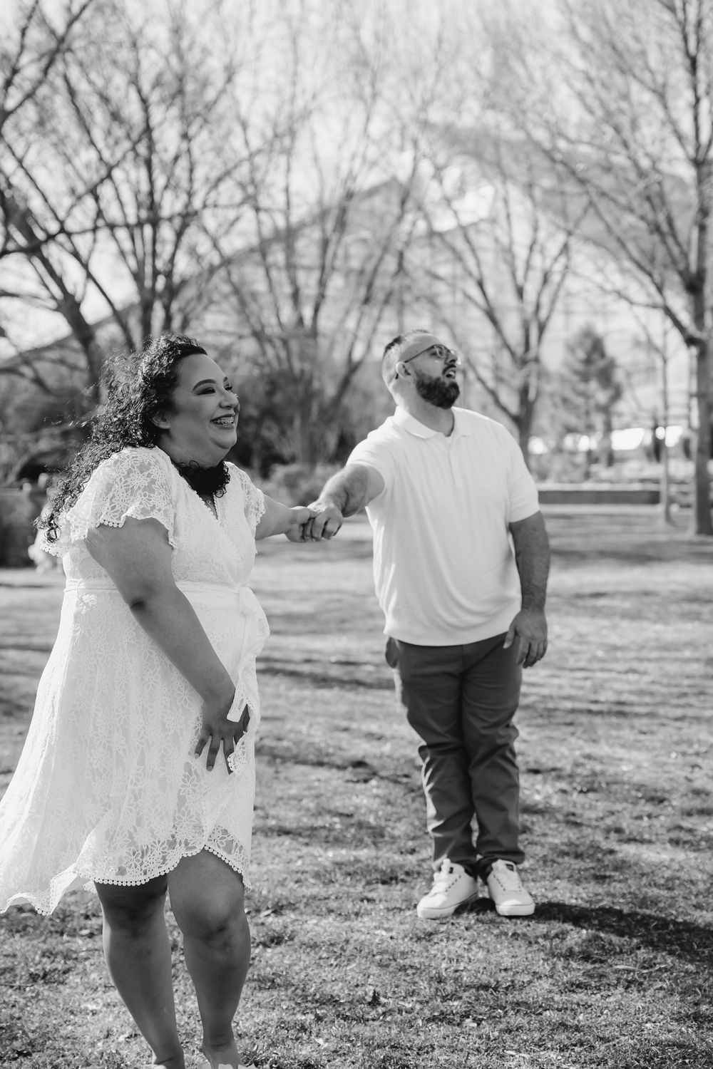 Black and white photo of a couple holding hands and walking in a park, woman in white lace dress smiling joyfully.