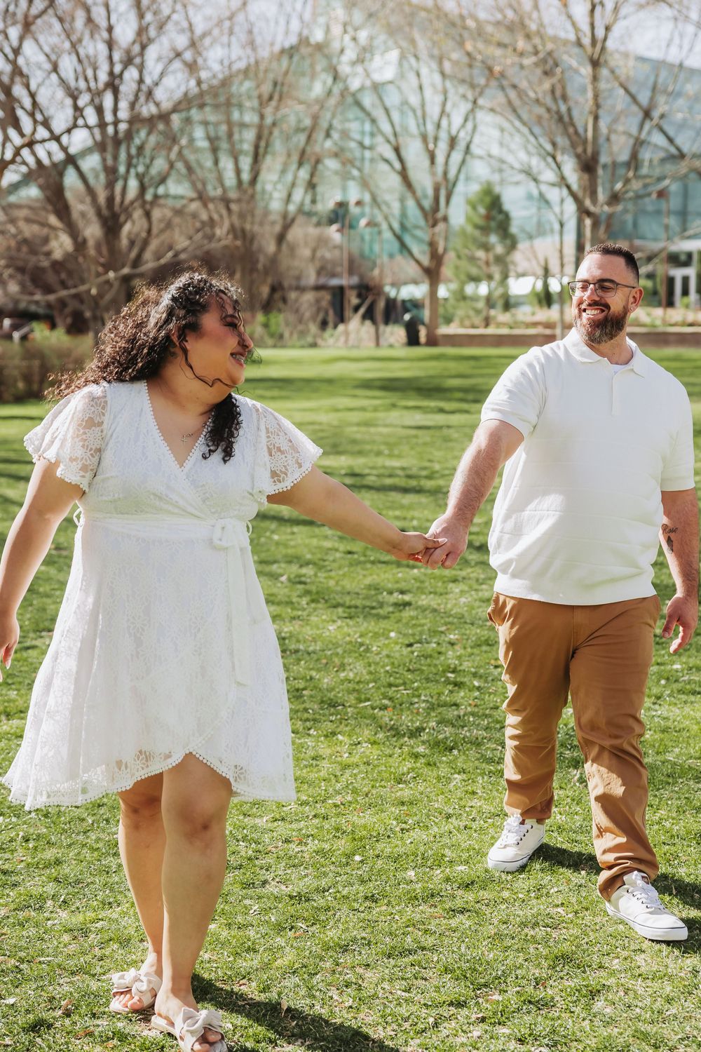 Couple holding hands and smiling while walking on green grass outdoors, woman in white dress, man in white shirt and tan pants.