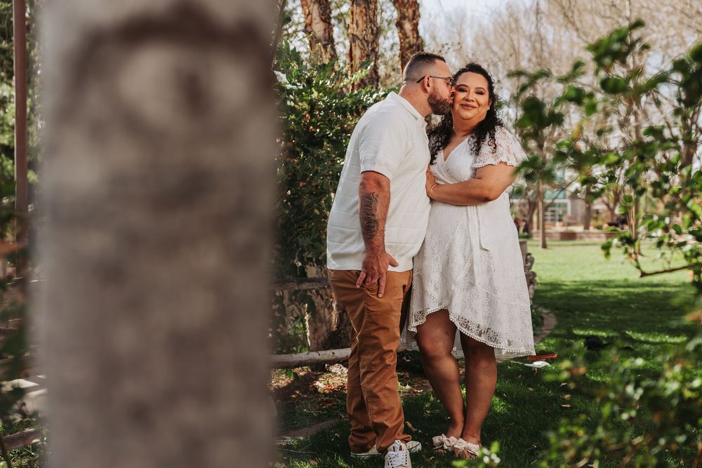 Couple sharing a tender kiss in a lush garden, man in white shirt and tan pants, woman in white dress.