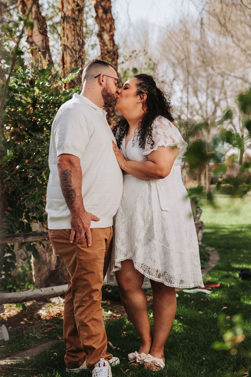 Couple sharing a kiss outdoors in a garden, man in white shirt and tan pants, woman in white lace dress.