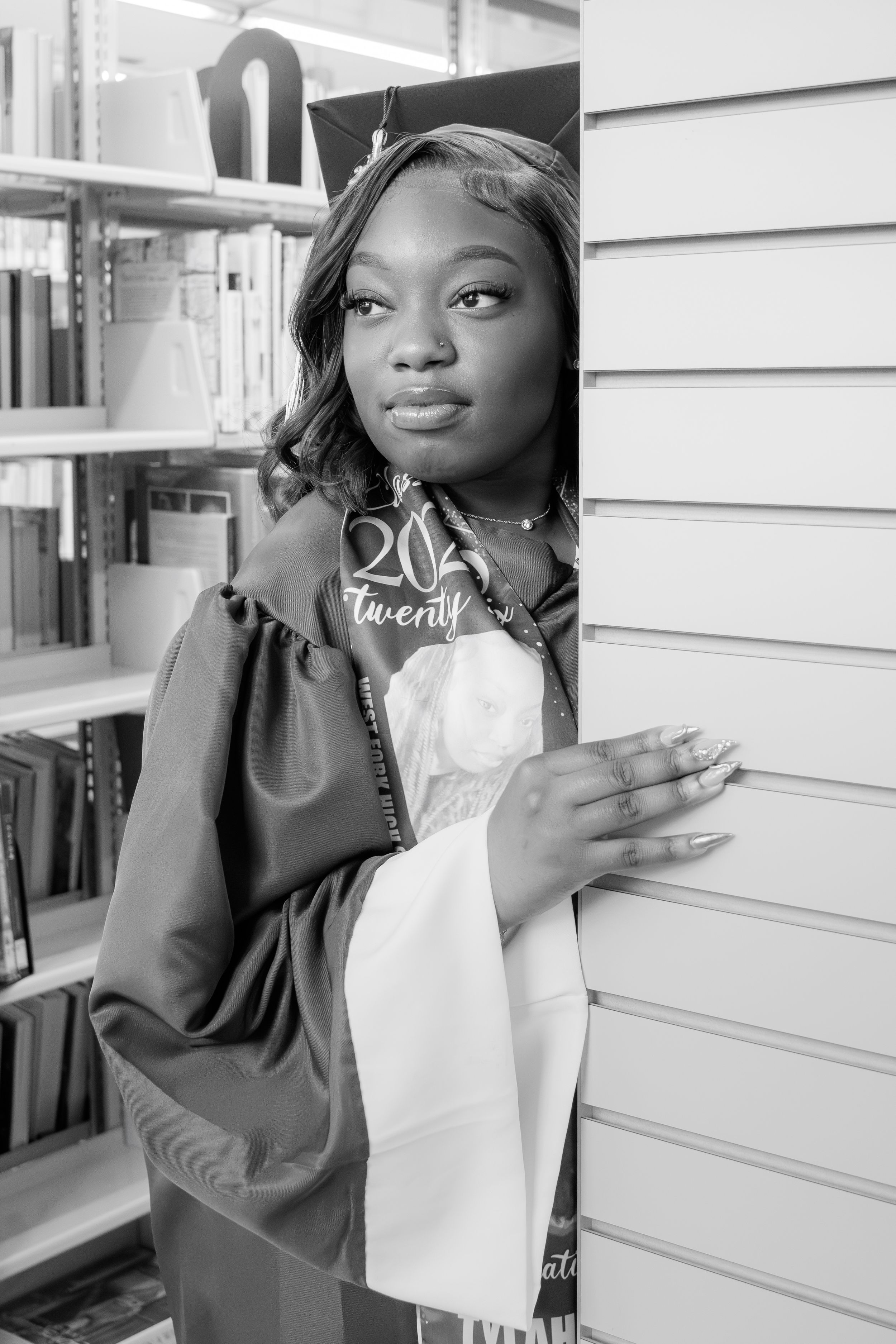 Black and white photo of a young woman in graduation cap and gown holding a diploma, posing in a library.