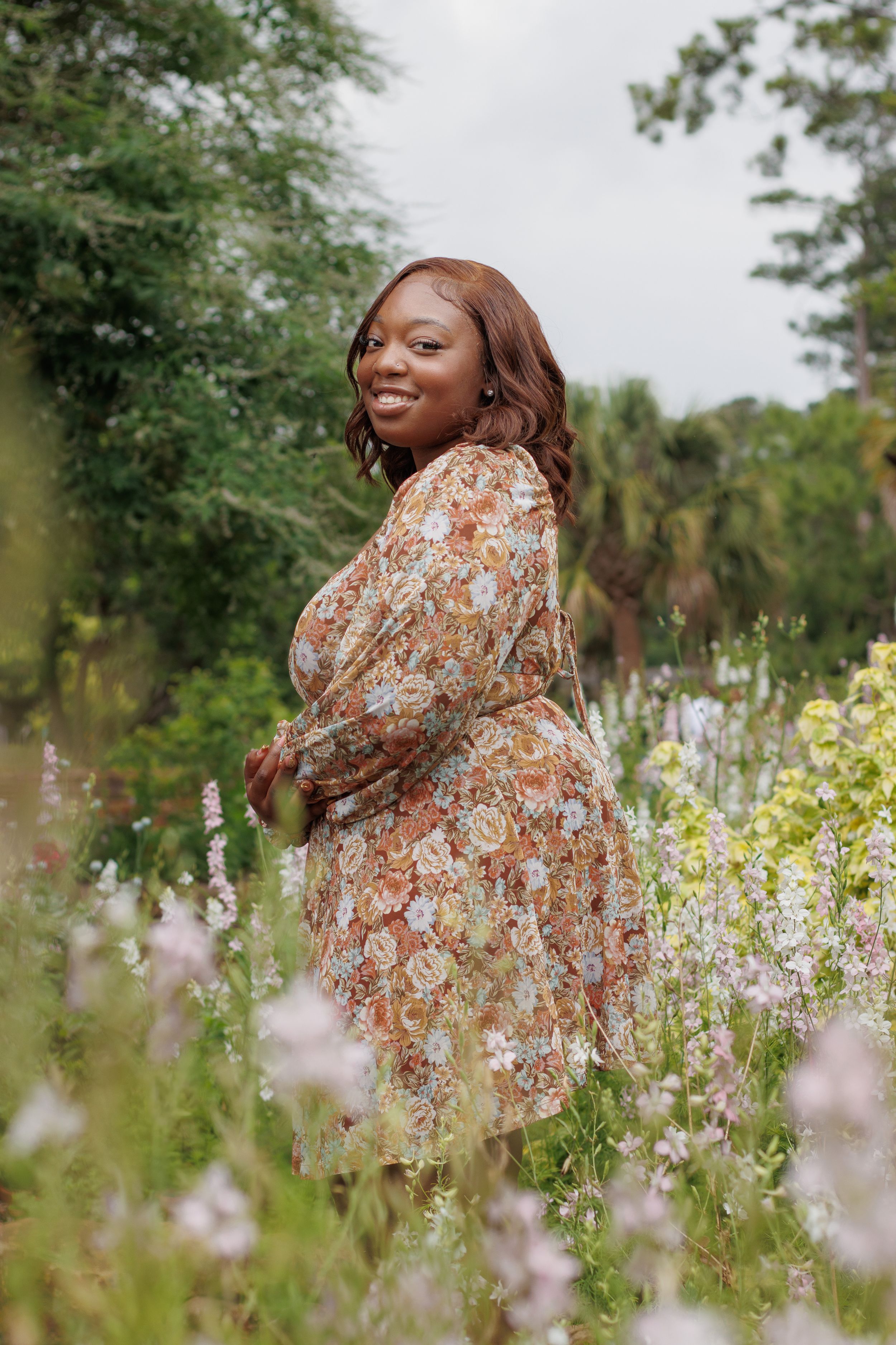 Smiling woman in floral dress standing among wildflowers in a lush garden setting.