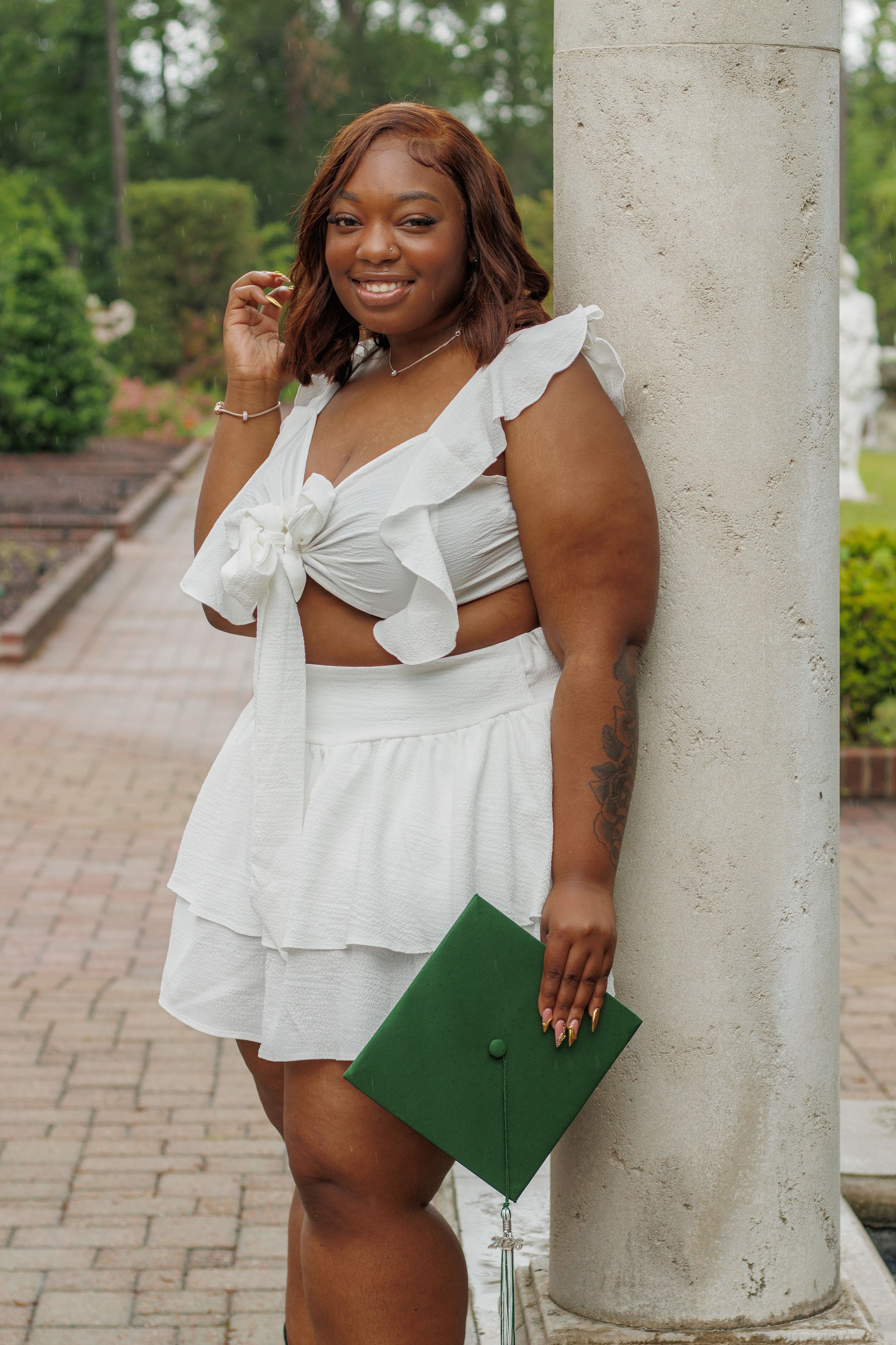 Smiling young woman in white ruffled crop top and skirt holding green graduation cap beside a column outdoors.